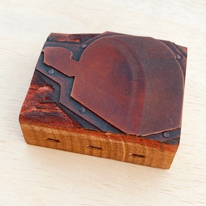 May include: A wooden printing block with a brown leather surface and metal accents. The block is rectangular and has a slightly rounded top. The leather surface is textured and has a worn look.