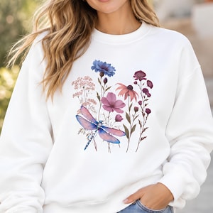 May include: A white crewneck sweatshirt featuring a central graphic print of a blue and purple dragonfly surrounded by various wildflowers in shades of pink, blue, and deep purple, including cornflowers and coneflowers.