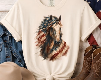 Puede incluir: Una camiseta de manga corta de color crema con un estampado gráfico de un caballo cuya crin y pelaje se mezclan con el patrón de la bandera estadounidense. La camiseta tiene cuello redondo, un dobladillo anudado y una etiqueta Comfort Colors.