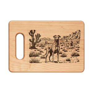 May include: A light-colored wooden cutting board with a handle. The board features a detailed engraved illustration of a dog standing in a desert landscape with cacti and mountains in the background.