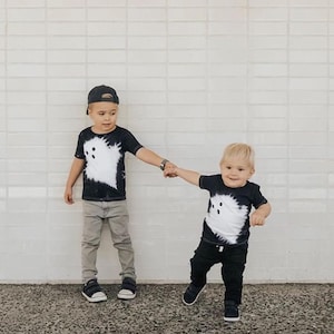 Peut inclure: Deux t-shirts noirs &agrave; manches courtes arborant un grand motif de fant&ocirc;me blanc de style tie-dye sur le devant. Les t-shirts sont port&eacute;s avec des pantalons d&eacute;contract&eacute;s et des baskets, pr&eacute;sentant un ensemble assorti &agrave; col rond.