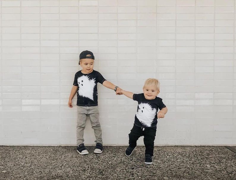 Peut inclure: Deux t-shirts noirs &agrave; manches courtes arborant un grand motif de fant&ocirc;me blanc de style tie-dye sur le devant. Les t-shirts sont port&eacute;s avec des pantalons d&eacute;contract&eacute;s et des baskets, pr&eacute;sentant un ensemble assorti &agrave; col rond.