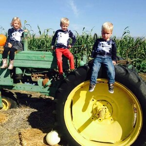 Peut inclure: Trois enfants portant des t-shirts &agrave; manches longues tie-dye assortis sont assis sur un tracteur vert vintage aux grandes roues jaunes. L'enfant de gauche porte une jupe sombre et des bottes, celui du centre un pantalon rouge et celui de droite un jean.