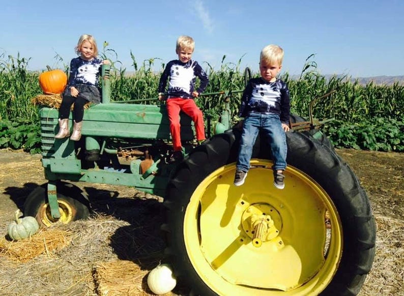 Peut inclure: Trois enfants portant des t-shirts &agrave; manches longues tie-dye assortis sont assis sur un tracteur vert vintage aux grandes roues jaunes. L'enfant de gauche porte une jupe sombre et des bottes, celui du centre un pantalon rouge et celui de droite un jean.