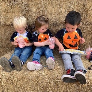 Peut inclure: Trois enfants portant des t-shirts noirs tie-dye avec des motifs de citrouille orange. Deux enfants tiennent des gobelets en plastique transparent avec des boissons roses et des pailles, tandis que le troisi&egrave;me tient un aliment envelopp&eacute; d'aluminium.
