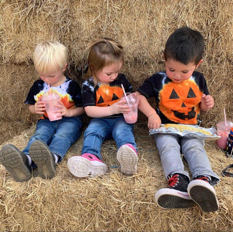 Peut inclure: Trois enfants portant des t-shirts noirs tie-dye avec des motifs de citrouille orange. Deux enfants tiennent des gobelets en plastique transparent avec des boissons roses et des pailles, tandis que le troisi&egrave;me tient un aliment envelopp&eacute; d'aluminium.