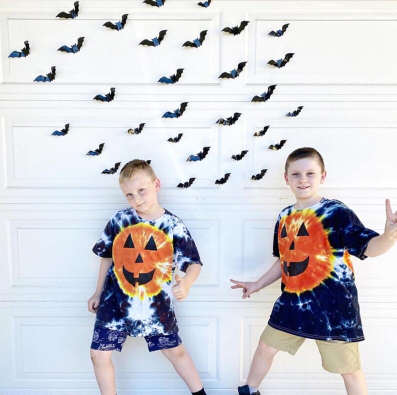 Peut inclure: Deux t-shirts &agrave; manches courtes tie-dye pr&eacute;sentant un grand motif de citrouille orange avec des yeux triangulaires noirs et une bouche dentel&eacute;e. Les t-shirts ont un motif en spirale noir, blanc et orange. De nombreuses petites chauves-souris noires sont fix&eacute;es au mur.
