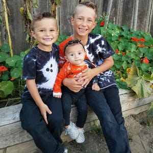 Peut inclure: Trois enfants assis ensemble. L'enfant &agrave; gauche porte un t-shirt tie-dye noir et blanc et un pantalon noir. L'enfant au centre porte un haut &agrave; manches longues orange avec un motif de toile d'araign&eacute;e, un legging noir et un bandeau noir. L'enfant &agrave; droite porte un t-shirt tie-dye noir et blanc et un pantalon noir.