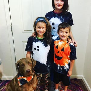Peut inclure: Trois enfants et un chien marron portant des v&ecirc;tements tie-dye. Deux enfants portent des t-shirts tie-dye noirs et blancs avec un motif de fant&ocirc;me, un enfant porte un t-shirt tie-dye orange et noir avec un motif de citrouille, et le chien porte un bandana assorti.
