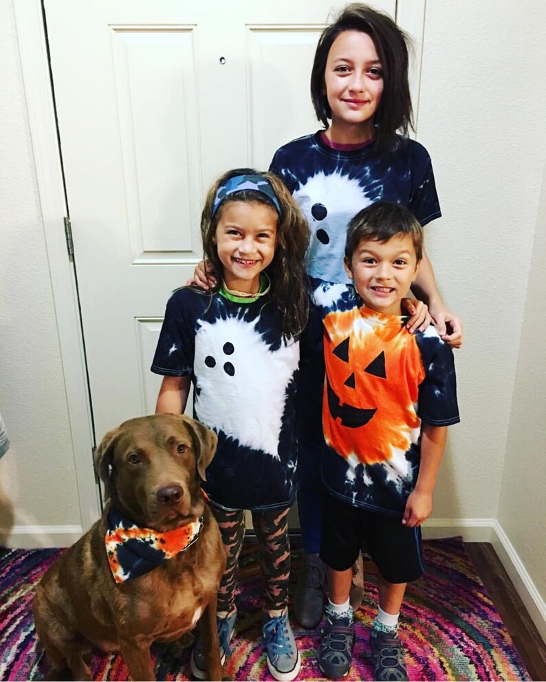 Peut inclure: Trois enfants et un chien marron portant des v&ecirc;tements tie-dye. Deux enfants portent des t-shirts tie-dye noirs et blancs avec un motif de fant&ocirc;me, un enfant porte un t-shirt tie-dye orange et noir avec un motif de citrouille, et le chien porte un bandana assorti.