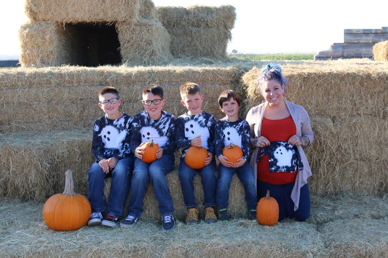Peut inclure: Quatre chemises &agrave; manches longues avec un motif tie-dye noir et blanc et un graphique de fant&ocirc;me blanc sur le devant. Les chemises sont port&eacute;es par quatre enfants et tenues par un adulte, accompagn&eacute;es de jeans bleus et de citrouilles orange.