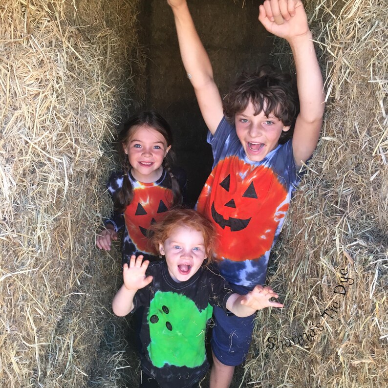 Peut inclure: Trois enfants portant des t-shirts tie-dye. Deux t-shirts pr&eacute;sentent des motifs de citrouille orange avec des traits noirs, et un t-shirt pr&eacute;sente un motif de fant&ocirc;me vert avec des traits noirs. Le texte Shamma's Tie Dye est visible en bas &agrave; droite.