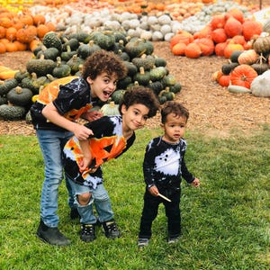 Peut inclure: Trois enfants portant des t-shirts tie-dye noir et blanc avec des motifs de citrouilles orange et des jeans en denim bleu ou noir. Les deux a&icirc;n&eacute;s portent des t-shirts avec de grandes citrouilles, le plus jeune a des d&eacute;tails de fant&ocirc;mes.