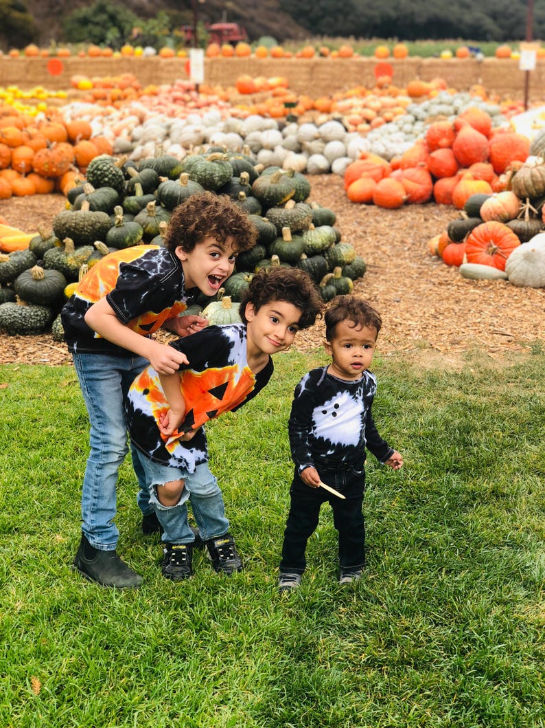 Peut inclure: Trois enfants portant des t-shirts tie-dye noir et blanc avec des motifs de citrouilles orange et des jeans en denim bleu ou noir. Les deux a&icirc;n&eacute;s portent des t-shirts avec de grandes citrouilles, le plus jeune a des d&eacute;tails de fant&ocirc;mes.