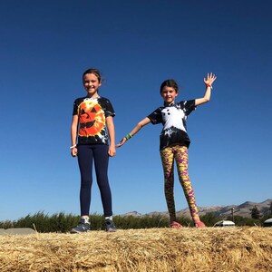 Peut inclure: Deux enfants debout sur une botte de foin. L'un porte un t-shirt tie-dye noir avec un motif de citrouille orange et un legging bleu marine. L'autre porte un t-shirt tie-dye noir avec un motif de fant&ocirc;me blanc et un legging jaune &agrave; motifs violets.