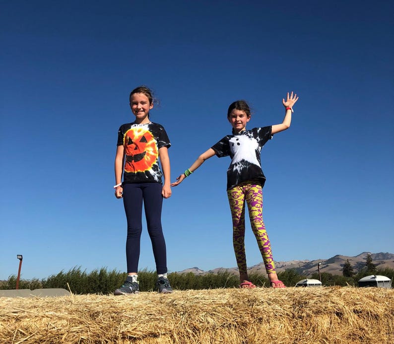 Peut inclure: Deux enfants debout sur une botte de foin. L'un porte un t-shirt tie-dye noir avec un motif de citrouille orange et un legging bleu marine. L'autre porte un t-shirt tie-dye noir avec un motif de fant&ocirc;me blanc et un legging jaune &agrave; motifs violets.