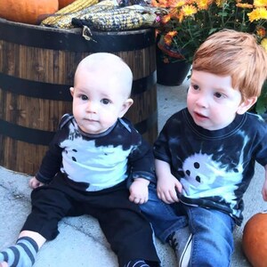 Peut inclure: Deux enfants portant des t-shirts tie-dye noir et blanc avec des motifs de fant&ocirc;mes. L'un porte un pantalon noir et des chaussettes ray&eacute;es, l'autre un jean bleu et des baskets grises. Les t-shirts pr&eacute;sentent un motif de fant&ocirc;me blanc central.