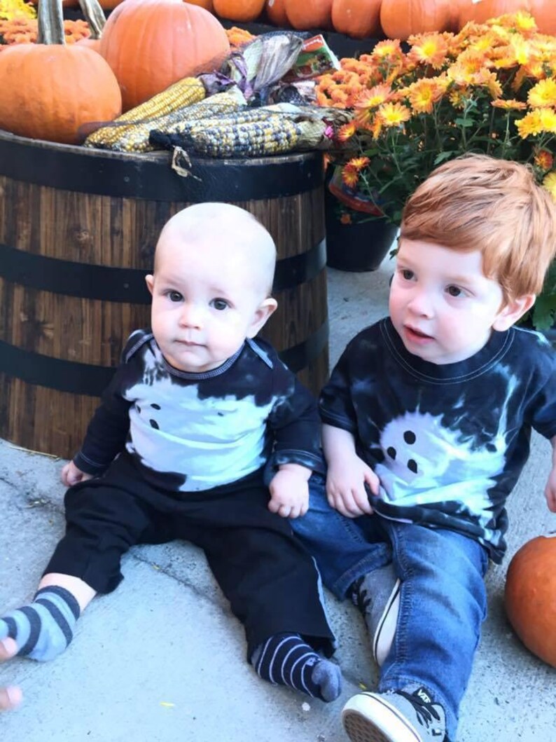 Peut inclure: Deux enfants portant des t-shirts tie-dye noir et blanc avec des motifs de fant&ocirc;mes. L'un porte un pantalon noir et des chaussettes ray&eacute;es, l'autre un jean bleu et des baskets grises. Les t-shirts pr&eacute;sentent un motif de fant&ocirc;me blanc central.