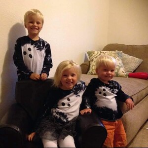 Peut inclure: Trois enfants portant des t-shirts &agrave; manches longues assortis avec un panneau central blanc orn&eacute; de trois points noirs et des manches &agrave; effet tie-dye noir. Les t-shirts ont une encolure ronde et une coupe ajust&eacute;e. Les enfants sourient.