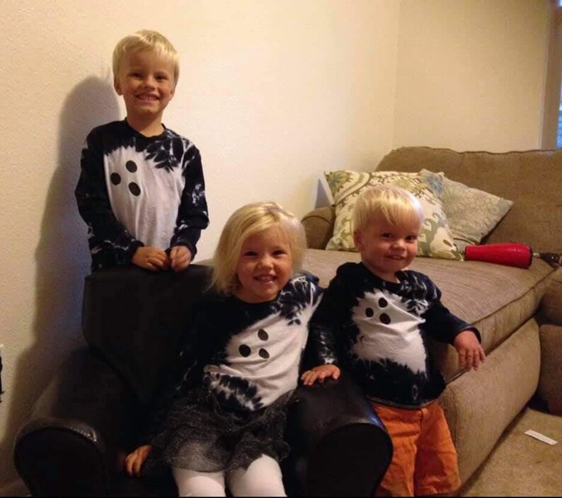 Peut inclure: Trois enfants portant des t-shirts &agrave; manches longues assortis avec un panneau central blanc orn&eacute; de trois points noirs et des manches &agrave; effet tie-dye noir. Les t-shirts ont une encolure ronde et une coupe ajust&eacute;e. Les enfants sourient.