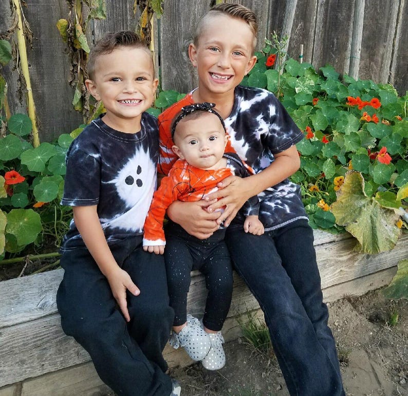 Peut inclure: Trois enfants assis ensemble. L'enfant &agrave; gauche porte un t-shirt tie-dye noir et blanc et un pantalon noir. L'enfant au centre porte un haut &agrave; manches longues orange avec un motif de toile d'araign&eacute;e, un legging noir et un bandeau noir. L'enfant &agrave; droite porte un t-shirt tie-dye noir et blanc et un pantalon noir.