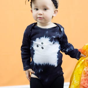 Peut inclure: Un body &agrave; manches longues noir avec un motif tie-dye, orn&eacute; d'un fant&ocirc;me blanc avec trois points noirs pour les yeux et la bouche, accompagn&eacute; d'un bandeau noir avec une grande fleur en tissu et d'un legging noir uni.