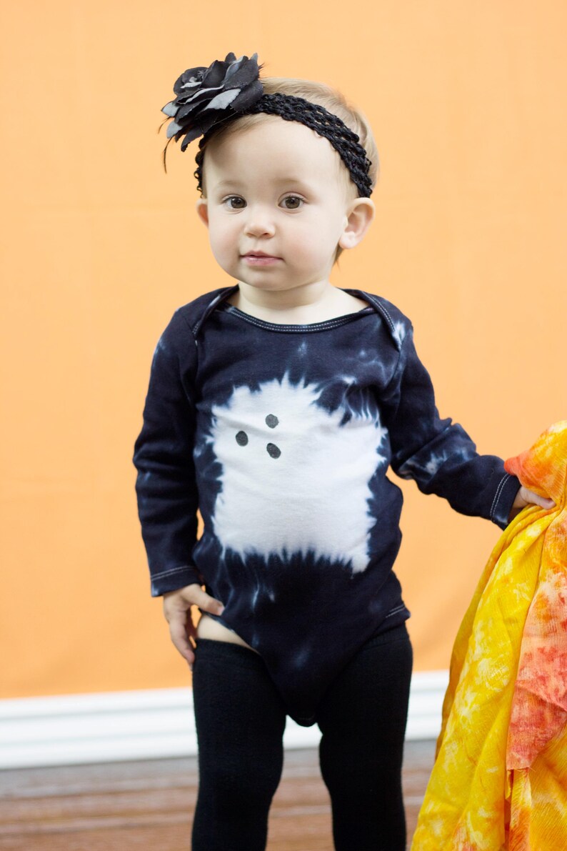 Peut inclure: Un body &agrave; manches longues noir avec un motif tie-dye, orn&eacute; d'un fant&ocirc;me blanc avec trois points noirs pour les yeux et la bouche, accompagn&eacute; d'un bandeau noir avec une grande fleur en tissu et d'un legging noir uni.