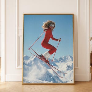 May include: Framed print of a skier in a red jumpsuit mid-air, skis angled downward. The skier wears goggles and white fur-trimmed boots. The background features snow-covered mountains and a clear blue sky. The print is in a gold frame.