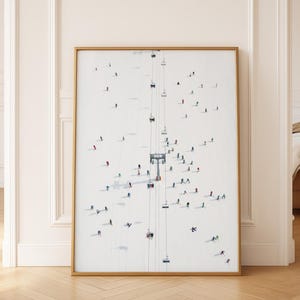 Può includere: Stampa artistica incorniciata raffigurante una scena sciistica invernale. L'opera mostra sciatori e una seggiovia su una montagna innevata. Gli sciatori indossano abiti colorati e la seggiovia è sospesa da cavi. La cornice è dorata.