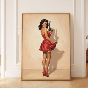 May include: A framed print depicting a retro illustration of a woman in a red dress holding a gun. The artwork has a vintage aesthetic, with the woman wearing red heels and stockings. The background is a neutral beige, and the frame is gold.