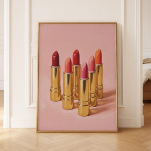 A framed retro lipstick poster featuring six golden lipsticks in shades of red, pink, and coral on a textured pink background, leaning against a cream wall in a bright, elegant interior with wooden floors and soft natural light. The artwork adds a feminine, vintage charm to the minimalist space.