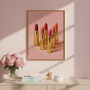 A framed retro lipstick poster featuring six golden lipsticks in red, pink, and coral shades on a pink background, hanging on a blush wall above a vanity with peonies, Chanel and Vogue books, and a perfume bottle. Elegant feminine wall art perfect for makeup rooms or fashion-inspired interiors.