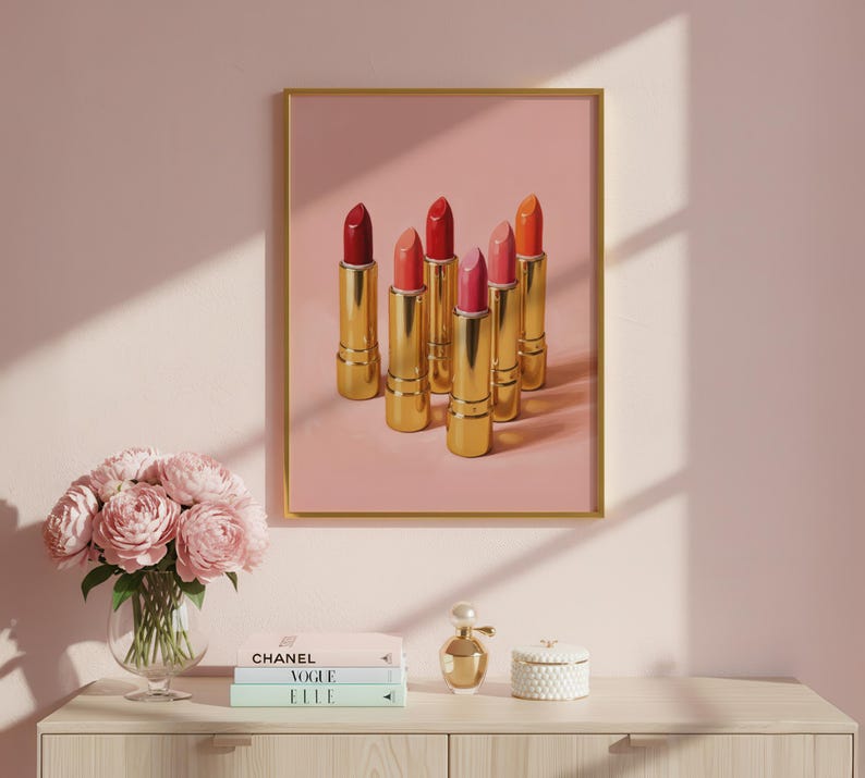 A framed retro lipstick poster featuring six golden lipsticks in red, pink, and coral shades on a pink background, hanging on a blush wall above a vanity with peonies, Chanel and Vogue books, and a perfume bottle. Elegant feminine wall art perfect for makeup rooms or fashion-inspired interiors.
