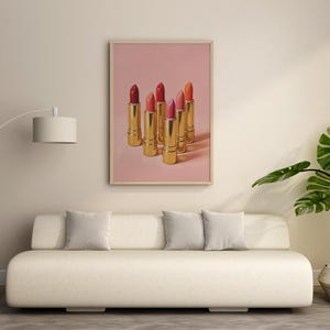 A framed retro lipstick poster featuring six gold lipsticks in red, pink, and coral tones on a soft pink background, hanging above a minimalist cream sofa in a modern living room. The decor includes a floor lamp, gray cushions, and a lush green plant, creating a stylish and elegant atmosphere with warm, natural light.