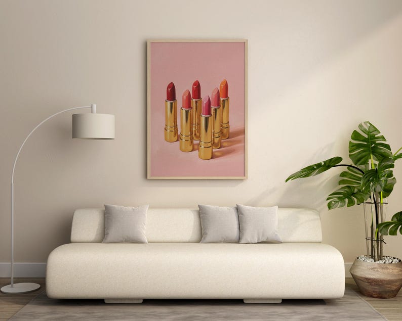 A framed retro lipstick poster featuring six gold lipsticks in red, pink, and coral tones on a soft pink background, hanging above a minimalist cream sofa in a modern living room. The decor includes a floor lamp, gray cushions, and a lush green plant, creating a stylish and elegant atmosphere with warm, natural light.