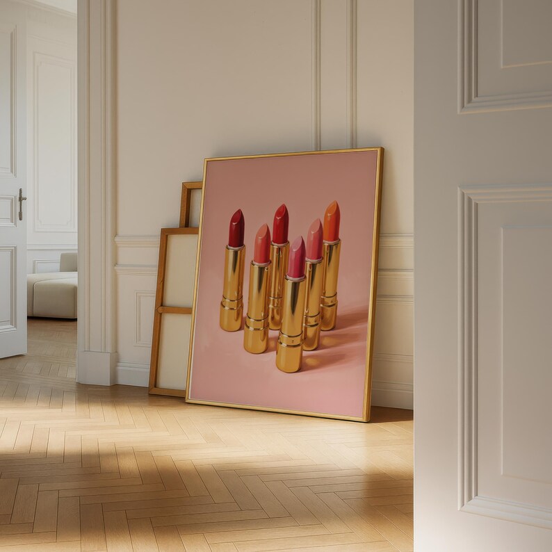 A framed retro lipstick poster featuring six gold lipsticks in red, pink, and coral hues on a pink background, leaning casually against a cream wall in a softly lit room with wooden floors and layered frames behind it. The warm light and elegant setting give the scene a refined, feminine, and vintage ambiance.