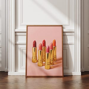 A framed retro lipstick poster featuring six golden lipsticks in red, coral, and pink tones on a pink background, leaning against a white paneled wall in a bright, elegant interior with wooden floors. Sunlight casts soft shadows, highlighting the artwork’s chic and feminine aesthetic.