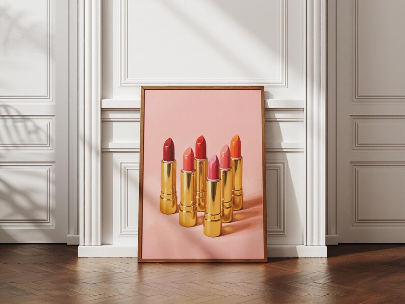A framed retro lipstick poster featuring six golden lipsticks in red, coral, and pink tones on a pink background, leaning against a white paneled wall in a bright, elegant interior with wooden floors. Sunlight casts soft shadows, highlighting the artwork’s chic and feminine aesthetic.