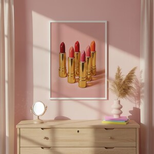 A framed retro lipstick poster featuring six golden lipsticks in shades of red, coral, and pink displayed on a pink background, hanging above a wooden vanity with soft sunlight, a small mirror, pampas grass in a vase, and pastel books. Perfect for a glam or feminine makeup room decor.