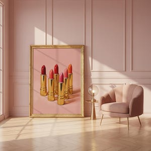 A framed retro lipstick poster featuring six golden lipsticks in shades of red, pink, and coral against a pink background, leaning on a blush wall beside a velvet armchair and gold lamp. The scene is bathed in soft sunlight, creating a luxurious, feminine, and retro-inspired atmosphere.