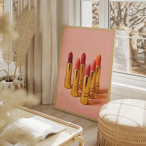A framed retro lipstick poster showcasing six gold lipsticks in shades of red, pink, and coral on a pink background, leaning against a bright window in a cozy, sunlit boho interior. The room features natural textures, beige tones, and soft décor elements like a woven basket, pampas grass, and a round ottoman, creating a warm, feminine atmosphere.