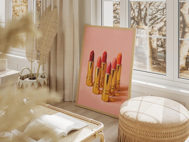 A framed retro lipstick poster showcasing six gold lipsticks in shades of red, pink, and coral on a pink background, leaning against a bright window in a cozy, sunlit boho interior. The room features natural textures, beige tones, and soft décor elements like a woven basket, pampas grass, and a round ottoman, creating a warm, feminine atmosphere.