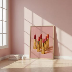 A framed retro lipstick poster showing six golden lipsticks in red, pink, and coral tones on a soft pink background, leaning against a blush wall beside stacked books and a small vase. Warm sunlight and minimalist decor create a cozy, feminine aesthetic