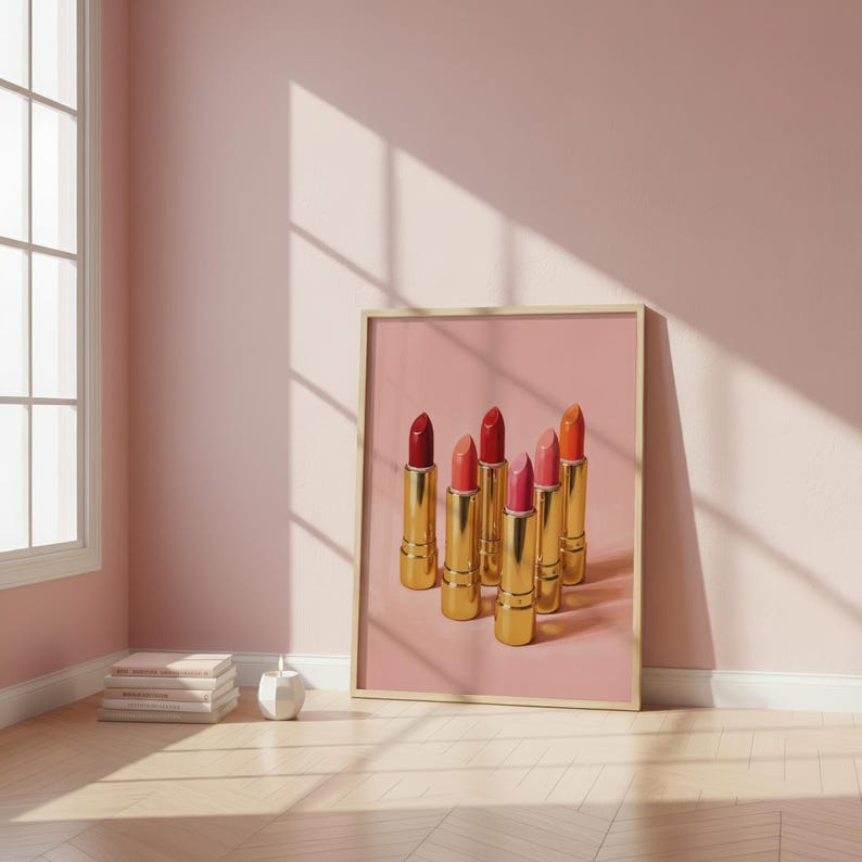 A framed retro lipstick poster showing six golden lipsticks in red, pink, and coral tones on a soft pink background, leaning against a blush wall beside stacked books and a small vase. Warm sunlight and minimalist decor create a cozy, feminine aesthetic