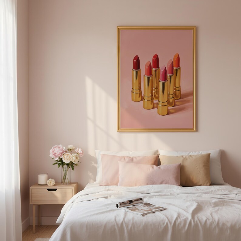 A gold-framed retro lipstick poster featuring six golden lipsticks in shades of red, pink, and coral displayed on a soft pink background, hanging above a neatly styled bed with pastel pillows and a vase of flowers on the nightstand. Elegant glam bedroom wall art with a feminine, vintage-inspired aesthetic.