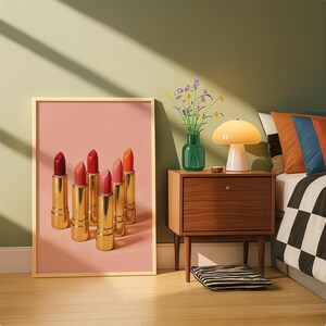 A framed retro lipstick poster featuring six golden lipsticks in red, pink, and coral tones on a pink background, leaning against a sage green wall beside a mid-century nightstand with a lamp and flowers. Warm sunlight and retro decor create a chic, stylish bedroom aesthetic.
