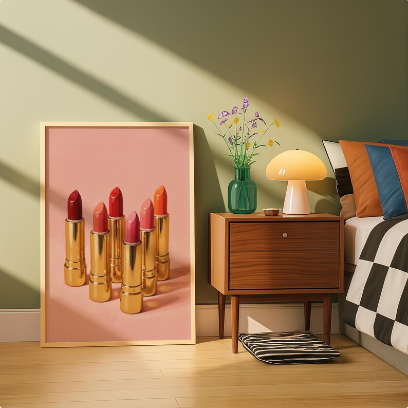 A framed retro lipstick poster featuring six golden lipsticks in red, pink, and coral tones on a pink background, leaning against a sage green wall beside a mid-century nightstand with a lamp and flowers. Warm sunlight and retro decor create a chic, stylish bedroom aesthetic.