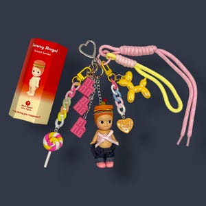 May include: A collection of Sonny Angel Snack Series charms and accessories. The charms include a figure with a hamburger hat, a lollipop, chocolate bar charms, a balloon dog, and a heart-shaped charm. The set is attached to a keychain with pink and yellow laces.