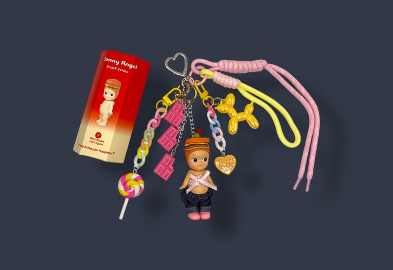May include: A collection of Sonny Angel Snack Series charms and accessories. The charms include a figure with a hamburger hat, a lollipop, chocolate bar charms, a balloon dog, and a heart-shaped charm. The set is attached to a keychain with pink and yellow laces.
