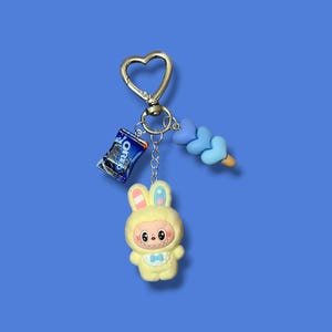 May include: A whimsical keychain featuring a yellow bunny character in a hooded outfit, a miniature Oreo cookie package, and a blue heart-shaped candy. The keychain has a gold-colored heart-shaped clasp and a blue background.